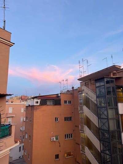 This image captures a dense urban architectural scene, viewed from an elevated perspective, likely during either sunrise or sunset. The sky, which fills the upper left portion of the frame (when oriented upright), displays a gradient from deep blue to a lighter blue, prominently featuring an elongated, soft pink and orange cloud streak.
The majority of the image is dominated by multi-story residential buildings, primarily on the right and bottom. Their facades are painted in warm terracotta, orange-brown, and lighter beige tones, characteristic of Mediterranean cities. Numerous windows of varying sizes are visible across the building exteriors; some are open, some closed, and a few display interior lights, suggesting evening hours or early morning activity. Several outdoor air conditioning units and various pipes or conduits are affixed to the walls.
Rooftops are densely populated with a multitude of television or radio antennas, indicating a long-established urban environment. One building, possibly a newer construction or renovation, features a distinct upper section composed of glass and dark metal framing, offering a contrast to the surrounding traditional stucco structures.
The scene conveys a quiet moment in a bustling city, likely Rome, Italy, given the architectural style and provided context. No people or specific events are visible. The interplay of the urban structures with the colorful twilight sky creates a sense of everyday life within a beautiful natural light. No visible text is present.