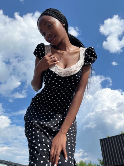 A young Black woman is posing outdoors against a bright blue sky with scattered white clouds. She is wearing a black polka dot dress with white lace trim around the neckline and puff sleeves. Her hair is styled in long braids. She is wearing a necklace and a ring on her left hand, which is placed on her chest. Her right hand is resting on her skirt. In the background, a dark, corrugated structure and some green foliage are visible, suggesting an urban or suburban setting. The image appears to have been taken from a low angle, looking upwards at the woman. The location in Kazan, Russia cannot be determined from the visual information provided.