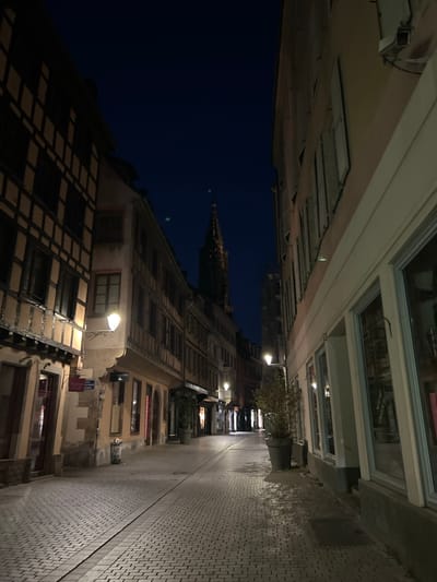 The image shows a street scene at night in Strasbourg, France, with a cobblestone path leading into the distance. The street is lined with historic buildings, some of which have timber-framed facades. Streetlights illuminate the scene, casting shadows on the cobblestones. In the background, a tall church spire is visible against the dark blue night sky. There are no people visible in the image. The overall impression is one of a quiet, atmospheric European city at night.