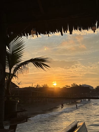 The image depicts a serene beach scene at sunset, likely in the city of Lhokseumawe, Indonesia. The sky is ablaze with warm hues of orange and yellow, characteristic of a tropical sunset, with scattered clouds catching the light. In the foreground, a thatched roof structure overhangs, with two simple hanging lights visible. To the left, the silhouette of a palm frond frames a portion of the view.
Below the structure, a sandy beach stretches along the coastline, with gentle waves lapping at the shore. Several small structures, possibly stalls or huts, are visible along the beach, supported by wooden posts. A few people are scattered along the water's edge and in the shallow sea; one individual walks along the wet sand, another stands in the water, and a third appears to be playing near the water.
In the mid-ground, a thick rope is strung horizontally, separating the immediate foreground from the beach area. A potted plant sits on a wooden surface in the lower left, partially obscuring the view of the beach. In the background, the silhouette of trees and distant buildings, including a dome-shaped structure, can be seen against the vibrant sky. The overall atmosphere is peaceful and tranquil, capturing the beauty of a coastal evening.