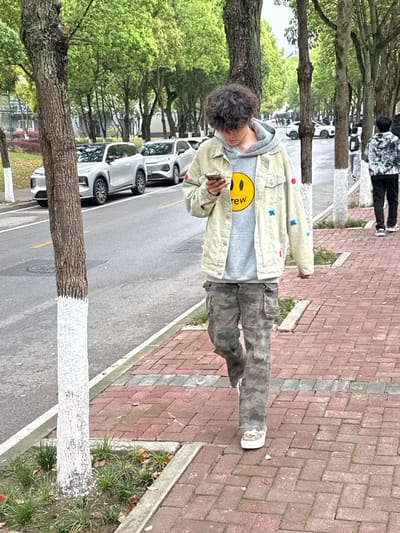 An outdoor scene in Jiangjin, China, depicts a person walking on a red brick sidewalk alongside a tree-lined street. The central figure, a young individual with dark, curly hair, walks from the right foreground towards the left, looking down at a light-colored smartphone held in both hands. They wear a light grey hooded sweatshirt with a yellow smiley face logo (Drew House brand), a light beige denim jacket adorned with pink and blue star-shaped patches, camouflage cargo pants, and white platform sneakers with leopard print accents. A white bandage or glove is visible on their left hand.

To the left of the main subject, another person in a black and white patterned hoodie and dark pants walks in the same direction on the sidewalk. Further down the street, several indistinct figures are visible. The asphalt road runs parallel to the sidewalk, marked with a yellow double centerline. Two light-colored SUVs are parked on the road, facing away from the camera; the closer vehicle has a dark roof.

Numerous trees with light green foliage line both the street and the sidewalk. Many tree trunks are painted white at their bases. Concrete curbs separate the sidewalk from planting strips containing sparse grass and fallen dry leaves, and from the paved road. The overall lighting suggests an overcast daytime condition.