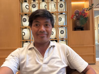 In a selfie taken in Kuta, Indonesia, a man with dark hair and brown eyes smiles at the camera. He is wearing a light-colored polo shirt. Behind him, a wooden shelving unit displays several white paper lanterns with black circular designs. To the right of the shelving unit, a vase of colorful flowers sits in a recessed alcove. The background appears to be an interior space with warm lighting and wooden accents.