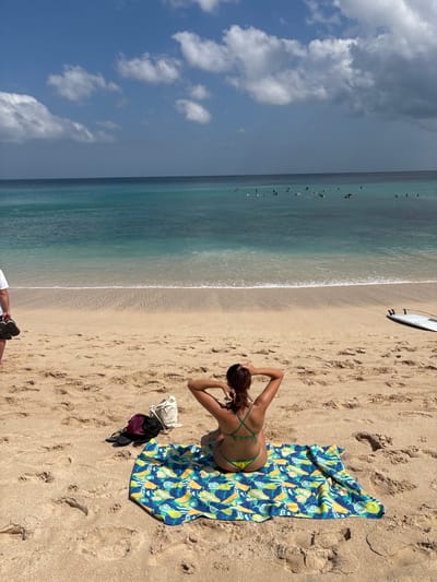The image depicts a beach scene with several people visible. In the foreground, a woman is lying on a colorful beach towel with a fruit pattern. She is wearing a green bikini and has her hands behind her head. Next to her are a white bag and a dark backpack. A white surfboard is lying on the sand further up the beach. In the ocean, there are several surfers scattered across the water. The sand is a light brown, and there are footprints visible. The water is a clear turquoise color, with white waves breaking near the shore. The sky is blue with some scattered white clouds, suggesting a sunny day. The lighting indicates it is likely daytime. The scene appears to be a popular beach destination, possibly in a tropical location, given the clear water and the presence of surfers. The overall mood seems relaxed and typical of a day spent at the beach. There is no visible text in the image.