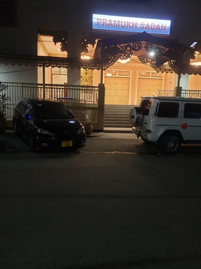 The image depicts the exterior of a building at night, identified by a prominent illuminated sign reading "PRAMUKH SADAN." The building features ornate architectural details, including decorative carvings above the entrance and intricate golden patterns on the facade. Two vehicles are parked in front of the building on an asphalt surface. To the left, a dark-colored minivan with a yellow license plate is visible. To the right, a white SUV, possibly a G-Wagen, is parked. The surrounding environment appears to be a street or parking area in an urban setting. The lighting suggests it is nighttime, with artificial illumination from the building's sign and internal lights, as well as possibly streetlights or external building lights, casting shadows. The scene does not contain any visible people. The setting is consistent with the city of Dar es Salaam, Tanzania, given the style of the building and the license plate format of the minivan.