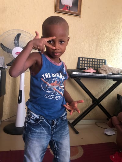 The image captures a young child standing indoors in Nkwelle Ezunaka, Nigeria. The child, of dark complexion and with short hair, is centrally positioned, looking directly at the viewer. The child's right hand is raised, with the index and middle fingers extended in a "V" shape near the face, while the left hand is held open to the side. The child wears a blue tank top adorned with a graphic of an airplane and the text "THE BIGGEST DUSTY," and faded blue denim jeans.
The background features a textured, light-yellow wall. To the child's left, a white standing fan with black blades and the "OX" logo is visible. To the child's right, an electronic keyboard rests on a black X-stand. Objects on the keyboard include a black grid-like component, a pink item, and light-colored crumpled material. Above the keyboard, a framed picture hangs on the wall, showing the lower portion of a person wearing white attire and red shoes. The floor consists of light-colored tiles, partially covered by a maroon rug with a circular pattern.