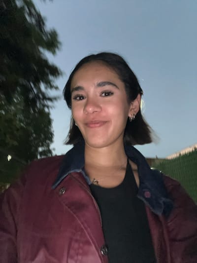 A medium shot of a young woman facing the camera. She has dark hair styled in a short bob, brown eyes, and is smiling slightly. She is wearing a black top and a maroon jacket with a blue denim collar. The photo appears to be taken outdoors in the evening, with trees and foliage visible in the background on the left and a blue-grey sky above. Based on the background details, the location may be San Andrés Cholula, Mexico. The photo appears to be taken from a slightly upward angle.
