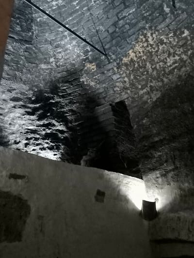 The image depicts the interior of a dimly lit, brick-lined structure, likely a cellar or an underground passage in Kuldīga, Latvia. The ceiling is a curved brick vault, showing signs of age and wear, with some areas appearing to be damp or discolored. A thin, dark cable runs diagonally across the ceiling.
In the lower part of the image, a rough, whitewashed wall extends horizontally. A light fixture, possibly a simple lamp or sconce, is mounted on this wall, casting a bright, localized glow that illuminates a section of the wall and creates shadows. To the right of the light fixture, a dark, cylindrical object, possibly a bucket or a piece of equipment, is partially visible.
Further into the structure, behind the illuminated wall, the brickwork of the vault continues, becoming darker and less distinct due to the limited light. There appears to be a darker, more enclosed area or alcove within the brick structure, possibly a recess or a passage. The overall impression is one of a historical, utilitarian space, possibly associated with old buildings or infrastructure in Kuldīga. There are no people or animals visible in the image.