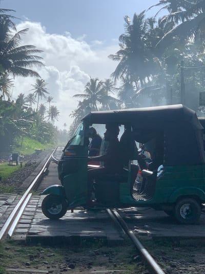 A green tuk-tuk, or auto-rickshaw, is parked on railroad tracks in Kodagoda, Sri Lanka. The tuk-tuk has a dark canvas roof and is occupied by at least two individuals, one of whom is driving and silhouetted against the bright sky. The vehicle is positioned on a crossing of the train tracks, which are made of steel rails laid on concrete sleepers. Surrounding the tracks, lush green vegetation, including numerous palm trees and banana plants, dominates the landscape. The sky is partly cloudy with bright sunlight filtering through, creating a hazy, atmospheric effect. In the distance, smoke or mist drifts through the trees. The scene suggests a tropical environment.