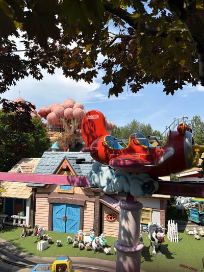 The image depicts a whimsical, cartoon-like outdoor scene, likely at an amusement park. The primary subject is a bright red amusement park ride designed to resemble an airplane. The ride features two rows of seats with blue interiors and yellow accents. The ride is adorned with "Fruit-tella" text. Scattered throughout the scene are various decorative elements that contribute to a playful, pastoral theme. These include several sheep figurines, some adorned with ribbons, and a cow figurine with black and white markings, wearing an orange bandana. A small blue toy truck is also visible. The setting includes stylized, colorful buildings with pitched roofs and wooden plank siding. One building features bright blue double doors. The ground is covered in artificial green turf. In the background, a roller coaster track can be seen, suggesting a larger amusement park setting. The sky is blue with some scattered clouds, indicating a clear, daytime scene. The overall atmosphere is cheerful and lighthearted, typical of a family entertainment venue. The image appears to be taken during the daytime, with the sun casting shadows. The presence of mature trees with dark green and reddish-brown leaves suggests it might be autumn. The location context provided is Castelnuovo del Garda, Italy.