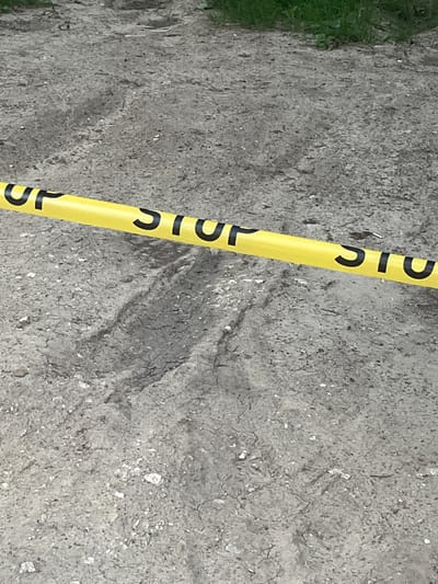 A close-up, eye-level shot shows a yellow caution tape with black lettering stretched diagonally across a dirt ground. The tape has the word "STOP" printed repeatedly in black letters. The ground is a mixture of dirt and small rocks, with visible tire tracks imprinted in it. In the upper part of the image, a patch of green grass can be seen. The scene appears to be outdoors, in an unpaved area. No specific landmarks or buildings are visible to determine a precise location within Chișinău, Moldova.