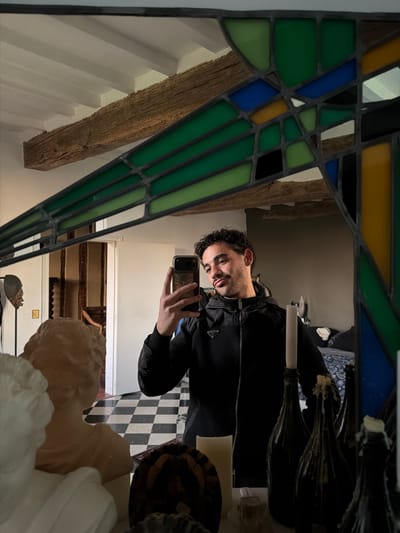 A male individual is observed indoors, taking a selfie reflection in a mirror within a building in Mauléon-d'Armagnac, France. The man has dark, curly hair, a mustache, and a short beard, and is wearing a black zippered jacket with a "PRADA" logo. He holds a smartphone with a gold-colored case in his right hand, pointed towards the mirror. The mirror is framed by leaded stained glass with geometric patterns in green, blue, yellow, black, and clear sections.
Reflected behind the individual, a room with a white ceiling featuring exposed, dark wooden beams is visible. A white doorway opens to an adjacent room, revealing a black and white checkered floor and further wooden architectural elements. To the man's left, a dark gray wall and a bed with a blue and white patterned duvet are reflected.
In the foreground, on a surface directly in front of the mirror, several objects are arranged. These include multiple dark-colored bottles, each fitted with a white candle, some displaying significant melted wax accumulation. A single tall, slender white candle stands upright. Two classical-style bust sculptures are also present; one is an earthy-toned bust showing the back of a head, and another partially visible white bust. A dark, textured, round object and a dark decorative object on a stand are also on the surface.