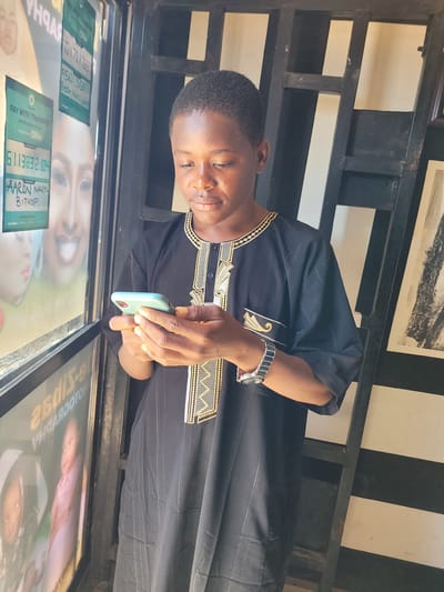 An upright, eye-level shot captures a young man indoors, likely in Jos, Nigeria, from the chest up. He has dark skin and short, dark hair, and is looking down intently at a light green smartphone held in his right hand. A silver-toned watch adorns his left wrist. He is dressed in a black tunic, featuring elaborate gold and black embroidery patterns around the neckline, on the upper chest, and on the sleeves, a style commonly seen in West Africa.
The background includes a dark, lattice-like partition or shelving unit, behind which a white wall and a framed picture are visible. In the foreground, below the man, is a reflective surface, likely a glass counter or window, displaying various advertisements and payment information.
Visible text on these displays indicates "PAY WITH TRANSFERS" options, suggesting a commercial transaction is taking place. One display shows "6113832104" for "AARON NAZILU BITKOP" via "Opay". Another offers "701 701 1667" and "P502500053" for "Opay", with the name "Fidelis Adom Nuhu" partially visible. The man appears focused on his phone, possibly facilitating a mobile payment or interacting with an application. The setting suggests an indoor commercial establishment during daytime.