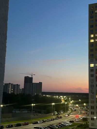 The image depicts an urban scene at dusk in Brovary, Ukraine. The sky transitions from a clear blue at the top to hues of pink and orange near the horizon, suggesting sunset. In the foreground, a portion of a light-colored building occupies the left edge, providing a frame for the wider view. To the right, a tall, multi-story yellow apartment building dominates the frame, with several windows illuminated from within.
Further into the scene, several other residential buildings are visible, some with lit windows, indicating occupancy. A construction crane stands prominently behind some of these buildings, suggesting ongoing development. A road curves through the middle ground, lined with streetlights, many of which are illuminated. Numerous parked cars are visible along the roadside and in parking areas, with a few vehicles in motion on the road, including one with its hazard lights on. The landscape beyond the road is characterized by trees and more distant buildings, fading into the twilight. The ground appears to be a mix of paved surfaces and green spaces. There are no people visible in the image.
