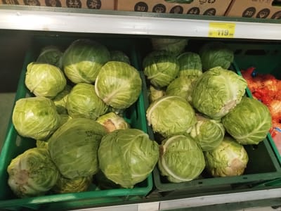 The image shows a retail display of fresh white cabbage heads in green plastic crates on a shelf in a supermarket. The cabbage is green and appears fresh. Some of the crates are full, while others are partially empty. In the background, there are boxes with Cyrillic text and images of vegetables. To the right of the cabbage, there are bags of onions. A yellow price tag with the text "119.99" is visible in the upper right portion of the image. The overall scene suggests a typical grocery store environment. The location is Yekaterinburg, Russia, as indicated by the Cyrillic text on the boxes.