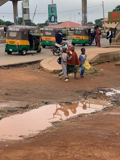 The image depicts a street scene in Gura Top, Nigeria, characterized by a large muddy puddle in the foreground. In the mid-ground, three young children are walking on the dirt path. The child on the left, wearing a grey and white patterned top and grey pants, is walking with their right arm raised. The middle child is wearing a brown headscarf and an orange top over a green and black checkered skirt. The child on the right is wearing a blue checkered shirt and blue pants, carrying a yellow bag. Behind the children, several auto-rickshaws, commonly known as 'keke napeps', are parked or moving. One auto-rickshaw, predominantly yellow and green, has the license plate "PTA-10760" visible. Another auto-rickshaw with the license plate "PTA-8926" is visible further back. A man is seated on a motorcycle in front of the latter vehicle. In the background, various buildings, including structures with red roofs and a building with a green sign featuring red lettering, are visible. A large billboard with an image of a man in a green shirt and the text "OPay 8 Shields" is also prominent. A fuel price display board for TotalEnergies is mounted on a pole near the auto-rickshaws. The overall environment appears to be an urban or semi-urban area with a mix of paved and unpaved surfaces, suggesting recent rainfall given the presence of the large puddle.