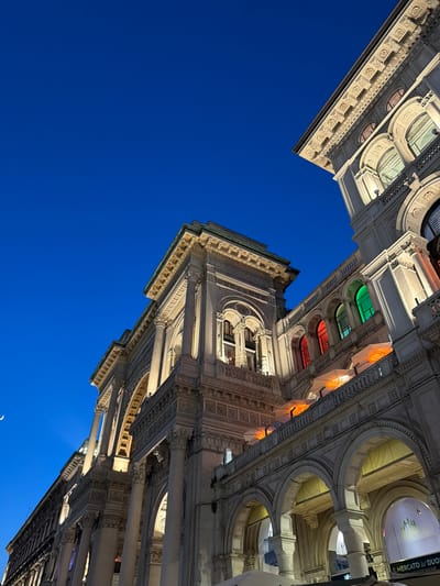 The image shows an upward-angle view of the Galleria Vittorio Emanuele II in Milan, Italy, at dusk. The architecture is grand and ornate, featuring classical columns, arched windows, and detailed stonework. Several windows are illuminated with colored lights: red, green, and orange are visible on one section of the building. The sky is a deep twilight blue, with a faint crescent moon visible in the upper left corner. Part of a sign reading "IL MERCATO DUO" is visible at the bottom right, suggesting commercial activity within the Galleria. The lighting from inside the Galleria highlights the intricate details of the facade. No people are clearly visible in the frame.