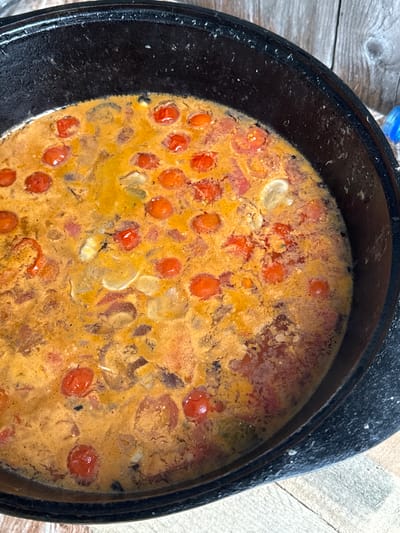 A black cast iron pot sits on a wooden surface, filled with a creamy, orange-hued stew containing numerous cherry tomatoes and sliced onions. The stew appears to be simmering, with small bubbles visible on the surface. The pot's rim is worn and shows signs of use. The background consists of weathered wooden planks. The image is taken from a top-down perspective. There is no information in the image to determine the location in the city of Sylva, Russia.
