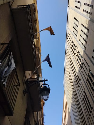 The image shows an upward view of buildings and flags in Barcelona, Spain. On the left, a building with balconies is visible, with a blue and white flag on the right balcony and a yellow and red striped flag on the left balcony. A lamp hangs from the left building. On the right, a tall, modern building with many small, rectangular windows is seen. The sky is clear blue.