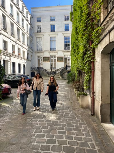 This image depicts a sunlit street scene in Paris, France, featuring three young women standing on a cobblestone street. The women are the central subjects, casually posed and looking towards the viewer. They are dressed in casual attire, including jeans, t-shirts, and light jackets, with one woman holding a phone.
The setting is characterized by classic Parisian architecture. To the left, a building facade is heavily draped with lush green ivy, contrasting with the stone structure. Behind the women, elegant, light-colored buildings with numerous windows and decorative wrought-iron railings line the street. A notable feature is a grand exterior staircase with ornate balustrades on one of the buildings. The ground is paved with traditional grey cobblestones. Several cars are parked along the street.
The time of day appears to be bright and clear, possibly morning or late afternoon, indicated by the visible blue sky and the soft shadows cast. The overall atmosphere is relaxed and pleasant, capturing a typical moment on a charming European street. No discernible text is visible on any of the buildings or objects.