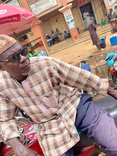 An eye-level, angled view depicts an African man wearing a brown and tan plaid shirt and navy pants, sitting on a red motorcycle. He wears sunglasses perched on his nose, and a tan hat, and his hands are visible on the motorcycle's handlebars. A building with "P.O.S" signs on it is in the background, with people sitting on the porch and inside the building. A red advertisement sign can be seen on the left side of the image. This scene is likely in Zaria, Nigeria, based on contextual clues, but that is not explicitly present in the image.