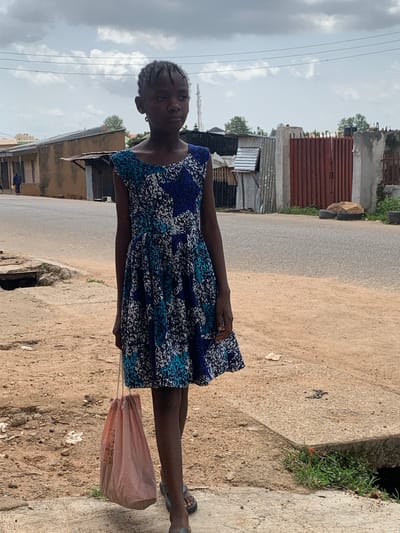 In Jos, Nigeria, a young Black girl stands on a dirt and concrete path next to a road. She is wearing a blue and white patterned dress and carrying a pink bag in her left hand. Her hair is braided and styled in small cornrows. In the background, there are buildings made of corrugated metal and concrete, some with red metal doors. A cell phone tower is visible in the distance. The sky is cloudy, with patches of blue peeking through. The ground is uneven and littered with debris.
