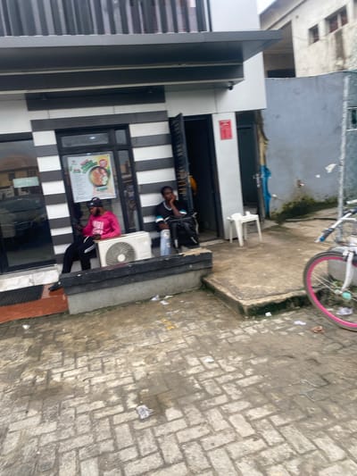 The image shows a street scene in Ado, Nigeria. A building with a modern facade featuring black and white stripes runs along the left side of the frame. Next to it, a person in a pink top and black pants sits with their back to the camera, facing an air conditioning unit mounted on the wall. Another person in a black shirt and blue jeans is visible further back, sitting with their legs crossed. A menu is displayed on the wall near the person in pink. In the background, a bicycle with a pink rim is propped up, suggesting it belongs to one of the individuals. The ground is paved with interlocking bricks and appears damp, possibly from recent rain. The overall atmosphere seems casual, with the people relaxed and taking a break. There are some scattered papers and debris on the ground, indicative of an urban street environment.