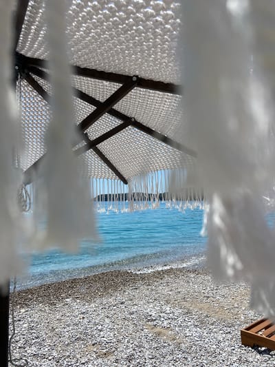 The image depicts a view of a beach and the sea, framed by a macrame umbrella with white tassels. The umbrella's canopy is made of white macrame material with a wooden frame. The tassels hang down, partially obscuring the view.

The beach is covered in small, grey and white pebbles. The sea is a vibrant turquoise blue, with gentle waves lapping at the shore. In the distance, a tree-covered peninsula or island is visible against a clear blue sky. To the right of the frame, a portion of a wooden beach lounger is visible amongst the pebbles.

The scene appears to be located in Prijevor, Montenegro, suggesting a coastal setting within this region. The lighting indicates it is daytime, likely sunny, given the bright illumination of the beach and sea. There are no people visible in the image. The focus is on the natural elements and the beach accessory.