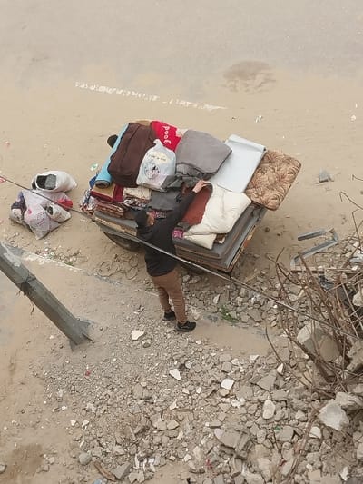 An overhead view captures an adult male interacting with a heavily laden utility vehicle or cart in Khan Yunis, Palestinian Territory. The male, dressed in a dark long-sleeved top, light brown trousers, and dark shoes, stands to the left of the vehicle's bed, reaching with his right hand to adjust or secure items. The vehicle bed is stacked with household goods, including a red blanket with "good" visible text, a gray blanket, a white patterned blanket, a dark brown bag, a plain white cushion, a floral-patterned cushion, a metallic panel, and several plastic bags, one partially displaying Arabic text. To the left of the vehicle, additional bundled items in white bags rest on the ground. The surrounding environment consists of unpaved, light brown earth extensively mixed with concrete rubble and scattered debris. A light-colored utility pole or beam is visible on the left side of the frame, embedded in the debris. To the right, a substantial pile of twisted rebar and other structural wreckage indicates a destroyed area. The background shows a broad expanse of light-colored, unpaved ground, likely a dirt road or open area.