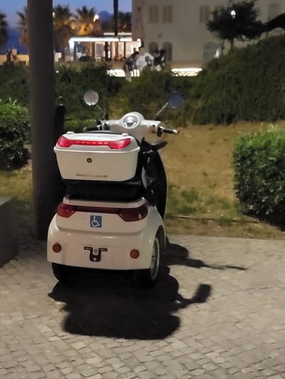A white three-wheeled scooter is parked on a cobblestone sidewalk at night. The scooter has a large white storage box on the back with red taillights and a blue wheelchair symbol sticker. There is a black pole to the left of the scooter. In the background, there are buildings with illuminated windows, green bushes, and palm trees. Several people are visible in the background, some walking and others standing. The scene appears to be in a city location in Quarteira, Portugal, possibly along the waterfront promenade.