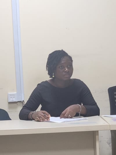 A medium shot captures a Black woman with braided hair sitting at a table in a room that appears to be an office or conference room. She is wearing a black long-sleeved shirt and has a pearl earring in her right ear. Her left hand rests on a stack of papers on the table, and she is wearing a ring on her left ring finger and a bracelet on her left wrist. She is looking to her right with a neutral expression. In the background, a pale wall is visible with a white electrical conduit and a white electrical outlet on the left. A sliver of a black office chair is visible on the right side of the frame. The table is a light beige color. The lighting is artificial and casts a subtle shadow. The scene is devoid of any specific cultural markers that would definitively place it in Abuja, Nigeria, or any other city.
