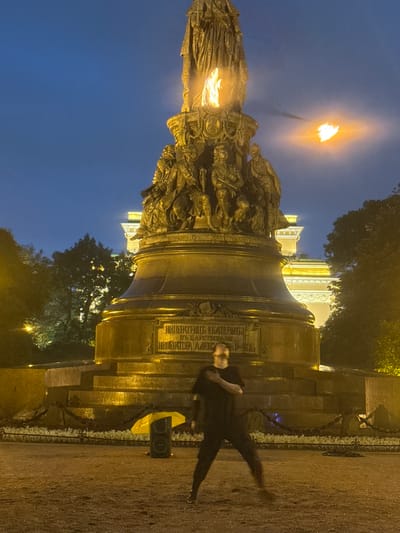 A person in dark clothing, blurred by motion, is captured in the foreground of the image. They are positioned in front of a large, ornate bronze monument, which is illuminated by flames at its top and on a torch extending from its side. The monument is detailed with sculpted figures and inscriptions. Behind the monument, a building with glowing yellow lights is visible. The scene is set at dusk or night, with a deep blue sky. The ground is a dirt or gravel surface. To the left of the monument, a dark speaker box sits on the ground, partially obscured by a bright yellow umbrella. The environment appears to be an outdoor public space, likely a park or square in Saint Petersburg, Russia, given the architectural style and the monument's appearance, which strongly resembles the Monument to Catherine the Great in Nevsky Prospekt.