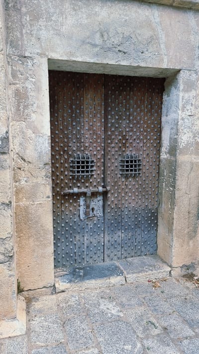 The image shows a heavy, weathered wooden double door set within a stone archway. The doors are studded with numerous small, square metal rivets. Each door has a small, square metal grate set into its upper half. A thick, horizontal metal bar slides through two latches mounted on the doors, securing them. One latch has a keyhole visible. The stone surrounding the door appears aged and textured, with some evidence of wear and chipping. The ground in front of the door is paved with rough cobblestones. The setting appears to be an exterior entryway. The location is the city of Montagut i Oix, Spain, specifically at the Church of Sant Pere, where this imposing door is a notable feature. There are no people or animals visible in the image.