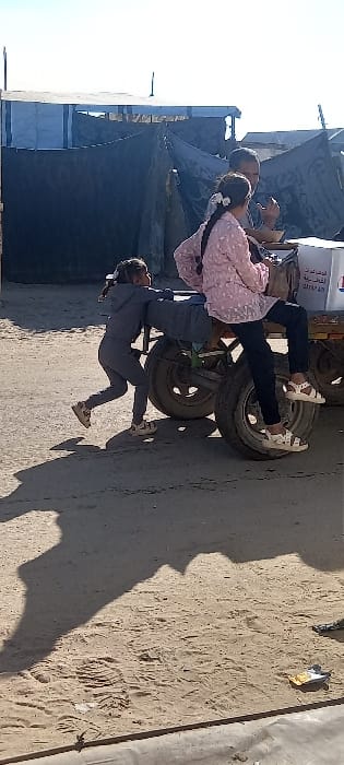 An outdoor scene in Khan Yunis, Palestinian Territory, depicts three individuals interacting with a two-wheeled cart on a dirt and sand surface under a clear sky. A young girl in a gray tracksuit and sandals is positioned on the left, actively pushing the cart from behind. Another girl, wearing a pink patterned long-sleeved top, dark pants, and white sandals, sits sideways on the cart's frame, with her legs dangling over one of the wheels. An adult male, partially visible behind the second girl, is also on or near the cart, appearing to hold something in his hand. The cart carries a white box with red and blue markings and Arabic text. In the background, several makeshift structures, constructed from tarpaulins and other materials, are visible, suggesting a temporary settlement. Long shadows are cast on the ground from the subjects and the cart, indicating direct sunlight.