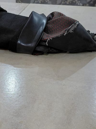 The image depicts a close-up, overhead view of a piece of fabric, possibly clothing or upholstery, lying on a tiled floor. The fabric is dark gray with a red and black striped pattern, and appears to be torn or frayed in places. A black, leather-like material is folded over a portion of the fabric. The floor is a light beige, textured tile. The scene is indoors and appears to be a dimly lit area. There are no people visible in the image. The location in the city of Santa Ana, Venezuela cannot be determined from the visual information provided.