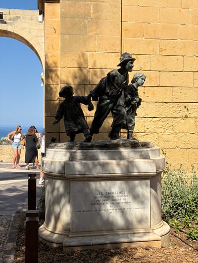 The image shows a bronze sculpture of three children walking, mounted on a marble pedestal in Valletta, Malta. The sculpture, titled "Les Cavroches," is by Maltese sculptor Antonio Sciortino and was presented to the government by the Society of Arts, Manufactures and Commerce in 1907. The pedestal has an inscription detailing this information.
The sculpture is positioned in front of a large, textured sandstone wall, with an arched opening to the left revealing a bright blue sky and the Mediterranean Sea in the distance. Several people are visible in the background, standing on a walkway to the left of the sculpture, some of whom appear to be tourists. The foreground features a cobbled pathway and a mulched garden area with some greenery. The lighting suggests it is daytime, with distinct shadows cast by the sculpture and the architectural elements.