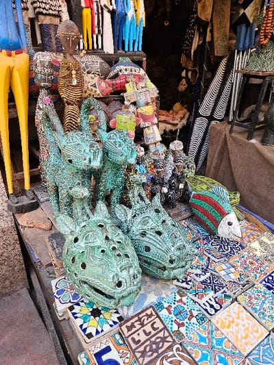 The image depicts a close-up view of an artisanal market stall in a souk within Marrakesh, Morocco. In the immediate foreground, two large, green-patinated metal sculptures of stylized animal or mythical creature heads, possibly dragons, rest on a surface. Each head features multiple circular perforations. Surrounding these heads and extending across the foreground are numerous hand-painted ceramic tiles of various sizes, exhibiting diverse geometric, floral, and calligraphic patterns in multiple colors. To the right, a ceramic fish sculpture painted with red, green, and white stripes is visible.
Behind the prominent metal heads, several smaller, green-patinated animal figures are arranged. The background is densely packed with a variety of handcrafted items. These include numerous tall, slender wooden figurines, some painted in bright primary colors such as blue, yellow, and red, representing stylized human forms. Other wooden carvings include a darker figure with a textured body and a carved face, and flat, painted wooden fish cutouts. Hanging textiles, some displaying black and white striped or green patterns, are visible in the upper and far-right background, delineating the rear of the stall. A dark opening suggests an interior shop space. No individuals are present within the frame.