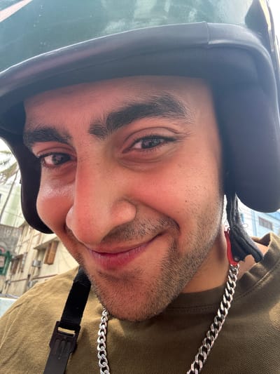 A close-up, eye-level, selfie-style, outdoor, daytime, medium shot captures a man wearing a green helmet and a brown shirt. The man is facing the camera and smiling. He has a silver chain around his neck. The background shows buildings and trees, suggesting an urban environment in Kolkata, India. The image is well-lit with natural light.