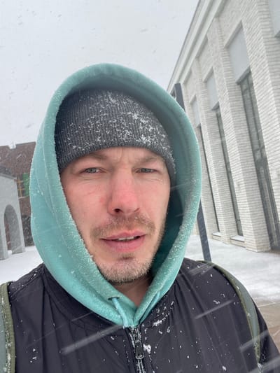 A man is facing the camera in a close-up shot with snow falling around him. He is wearing a grey beanie under a light green hooded sweatshirt. The right side of his body is visible, and he is wearing a black jacket with a zipper. The background reveals an urban environment with a light-colored building with a multi-layered facade. The man is positioned outside in the city of Chemoshur, Russia during snowfall.