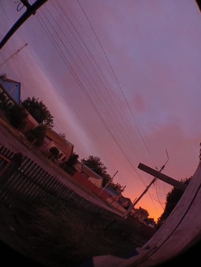 The image is a wide-angle, fisheye lens photograph of a sunset in Krasnyye Barrikady, Russia. The sky is a gradient of purple and orange, indicating the setting sun. Power lines and poles crisscross the sky. In the foreground, a wooden fence runs along the bottom left of the frame, with grass and some vegetation behind it. The middle ground shows a street with houses and trees. The houses appear to be of various sizes and colors, with some having red roofs. The overall impression is that of a rural or suburban setting during twilight. There are no people visible in the image.