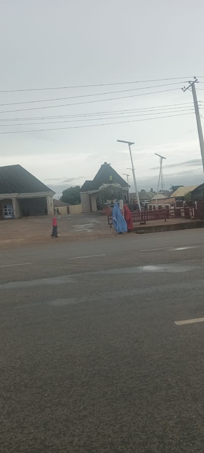 The image depicts a scene in Lafia, Nigeria, likely near a building with a gated entrance and what seems to be a cart selling items. Two people draped in blue and red garments are near the gate. Another person, wearing a red head covering, walks in the vicinity. Modern streetlights are in place. Power lines are visible above. The asphalt road in the foreground shows signs of minor water puddles. The sky is overcast.