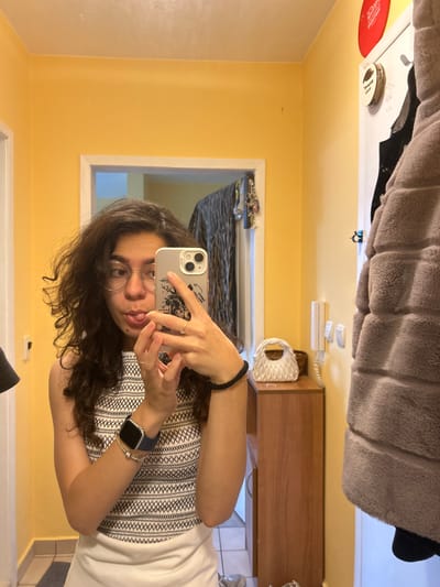 The image shows a young woman with curly dark hair and glasses taking a mirror selfie in what appears to be a home hallway or entryway. She is wearing a white and black patterned top and has her tongue sticking out playfully. She is holding a smartphone with a case featuring a sketch and her left hand is near her face. On her left wrist, she wears an Apple Watch and a silver bracelet. A dark-colored bracelet is visible on her right wrist.

The environment includes a yellow painted wall, a white door frame, and a wooden cabinet with a white decorative object and a potted plant on top. A grey furry coat is hanging on the right side of the frame, obscuring part of the door and wall. A white intercom system is mounted on the wall next to the cabinet. To the left of the woman, a glimpse of another door frame and a striped rug on the floor can be seen. On the wall above the door frame, a red circular object is visible. The lighting suggests an indoor setting, possibly in the evening or on a cloudy day. The scene does not provide any specific clues to pinpoint a location in Sofia, Bulgaria.