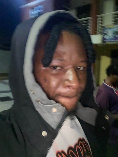 A close-up, tilted, and slightly blurry selfie shows a young Black person wearing a black jacket with a grey hood lining and a white shirt with orange lettering. The person's face is the central focus, with their dark hair framing their forehead and eyes. Their skin appears shiny, possibly from sweat or oil. The background is out of focus, but hints of indoor elements like shelving and possibly other people are visible. The lighting suggests it is nighttime or indoors with artificial light. The person's expression is neutral. No visible text is present in the image. The provided location context of Gura Top, Nigeria, is noted.