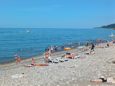 This media file depicts a bustling beach scene on a sunny day in Sochi, Russia. The image is filled with people enjoying a summer vacation. On the pebble beach, individuals are sunbathing on lounge chairs and towels, while others are wading in the clear blue water. Children are visible playing in the shallow parts of the sea.
Further out in the water, a white boat with an inscription of "Victoria" on its side is moving, creating a wake. Several people are swimming or floating in the sea, and some are even on paddleboards. The overall atmosphere appears relaxed and joyful, with many people engaging in typical beach activities.
The sky is a vibrant, clear blue, indicating good weather, and the sun is high, suggesting it is midday. The background shows a lush green hillside sloping down towards the coast, a common feature of the Sochi landscape. The sea stretches out to the horizon, meeting the sky in a clean, sharp line. There are also some buoys or markers visible in the water, possibly indicating swimming zones. The image is rich with detail, capturing the essence of a lively summer day at the seaside.