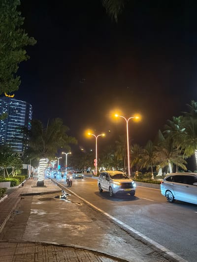 This media file depicts a nighttime scene on a city street, likely in Nha Trang, Vietnam, characterized by warm street lighting and illuminated buildings. The primary subjects include several cars and many motorcycles on the road, with a few pedestrians visible on the sidewalk. The scene is set along a street lined with palm trees, some of which are decorated with string lights. A modern building with vertical white lights dominates the background on the left.
The activity appears to be typical evening street life, with vehicles moving and people walking. The time of day is clearly night due to the dark sky and artificial lighting. The weather seems to be clear, as there are no indications of rain or fog. The presence of palm trees and the architectural style of the building suggest a tropical or subtropical coastal city.
Notable details include the distinctive orange glow of the streetlights, the reflections on the wet pavement, and the decorative lights adorning the palm trees and the building. The overall mood is lively yet relaxed, typical of a city street at night. No visible text is discernible in the image.