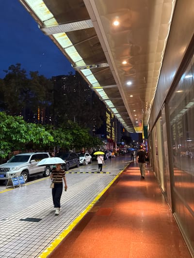 This image depicts a street scene in Tianhe District, China, at dusk or night. A covered walkway with reflective panels and overhead lighting extends along the right side of the frame, casting a warm orange glow on the wet pavement below. To the left, a sidewalk with a checkered pattern is bordered by a yellow stripe and leads to a street with parked cars. Beyond the street, lush green trees and tall buildings with illuminated windows are visible against the dark blue sky. Several people are walking under umbrellas, indicating it is raining. One person, a woman with dark hair, is walking towards the viewer under a light-colored umbrella, wearing a striped shirt and dark pants. Another person, a man with dark hair, is walking away from the viewer further down the covered walkway, wearing a dark t-shirt and light-colored pants. Other individuals and vehicles are present in the background, contributing to the bustling urban atmosphere.