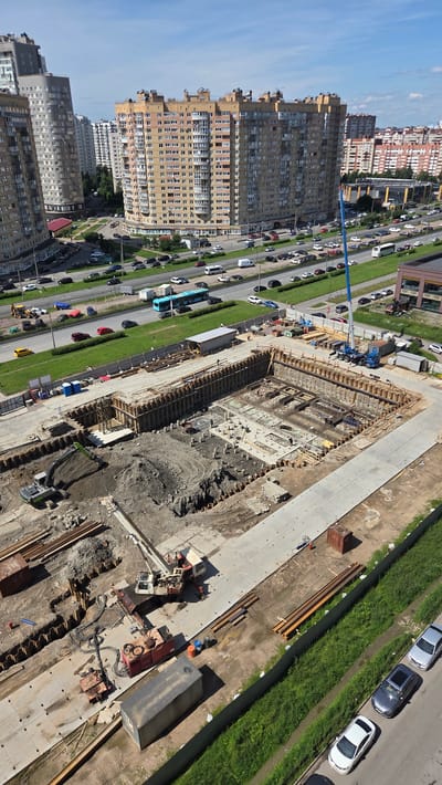 This aerial photograph captures an urban construction site in Saint Petersburg, Russia, on a clear, sunny day. The scene is dominated by the ongoing construction of a large building, with foundations and structural elements visible. A blue boom lift and various construction vehicles, including an excavator and a crane, are present on-site, indicating active work.
Adjacent to the construction zone, a multi-lane road is bustling with traffic, featuring a variety of cars and a blue bus. The road is bordered by a strip of green grass and mature trees. On the left side of the image, a row of tall, multi-story apartment buildings stands, typical of urban residential areas. The buildings vary in architectural style and color, with one prominent beige building exhibiting a complex facade with numerous balconies.
The time of day appears to be daytime, suggested by the bright sunlight and clear blue sky, casting distinct shadows on the ground. The overall impression is one of urban development and daily life coexisting with industrial activity. No text is clearly legible in the image.