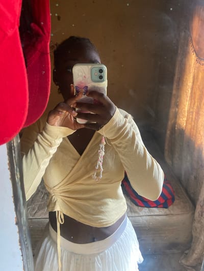 A person is taking a selfie in a mirror. They are holding a phone with a butterfly case. They are wearing a light yellow long-sleeved top and a white skirt. In the background, a mattress with a red and blue striped blanket is visible. The scene appears to be indoors, possibly in a bedroom. The lighting suggests it might be daytime.