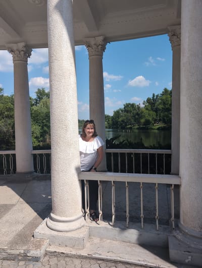 A woman is leaning over a white railing, looking at the camera. She is wearing a white shirt and has sunglasses on her head. She appears to be in a white gazebo with columns. Through the gazebo, a lake and trees can be seen. The sky is blue with some white clouds. The overall impression is a sunny day, likely in the late morning or early afternoon. The location is Kryvyi Rih, Ukraine.