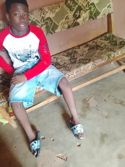 A young Black male is seated on a wooden bench with a patterned cushion. He is wearing a red and white long-sleeved shirt and light blue denim shorts. His feet are adorned with black slides featuring a white pattern. The bench is positioned on a concrete floor that appears worn and uneven, with some debris scattered around. The background suggests an indoor or covered outdoor setting, possibly in Akwanga, Nigeria, characterized by the simple furnishings and flooring.