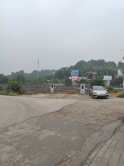 The image, taken on a hazy day, depicts a rural roadside scene in Jianyang City, China, featuring an electric vehicle charging station. In the foreground, a silver electric car, identified by the license plate "川A B85063", is parked next to a white charging station. Behind the car and charging station, a blue sign with white lettering indicates "永发停车场" (Yongfa Parking Lot) and "住房出租" (Housing for Rent), along with phone numbers. A second, identical charging station is visible to the left, further back and partially obscured by foliage. The surrounding environment consists of a dirt and gravel area leading to a paved road, with a large area covered by a mesh structure, possibly a greenhouse or crop protection, extending into the background. Lush green trees and rolling hills form the backdrop, with a telecommunications tower and power lines visible in the distance. The overall atmosphere is overcast and slightly subdued.